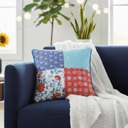 Colorful patterned pillow on a blue couch with a white blanket and plants in the background.