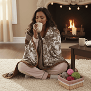 Woman sitting on the floor wrapped in a blanket, drinking from a mug in a cozy living room with a fireplace.