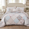 A patchwork quilted comforter set in a bedroom, featuring light blue, pink floral, and striped diamond panels with a ruffled edge, styled on a bed with a distressed cream headboard and matching nightstands. 