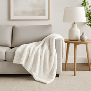 White faux fur white blanket with sherpa draped over a gray sofa in a living room with a lamp and side table.
